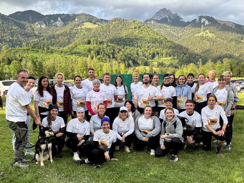 Eine große Gruppe von Menschen in weißen T-Shirts posiert lächelnd für ein Gruppenfoto auf einer Wiese vor einer Bergkulisse. Links im Vordergrund sitzt ein Hund.