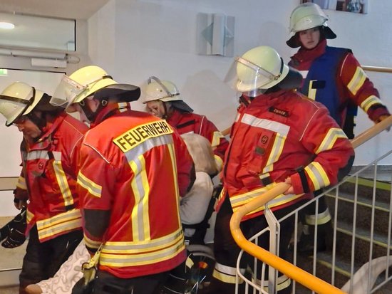 Feuerwehrleute in roter Schutzausrüstung und Helmen tragen eine Person die Treppe hinunter. Auf dem Rücken eines Einsatzmannes ist deutlich „FEUERWEHR“ zu lesen. Die Gruppe bewegt sich an einem orangefarbenen Geländer entlang, vermutlich bei einer Rettungsübung.