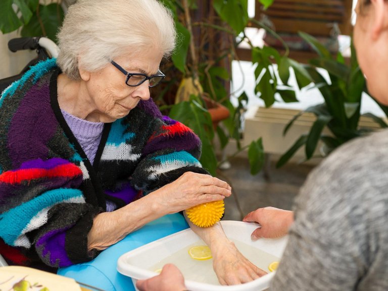 eine alte Frau hält eine Hand in eine Schüssel mit Wasser (und Zitronenscheiben). Dabei rollt sie mit einem Gummiball über ihren Arm