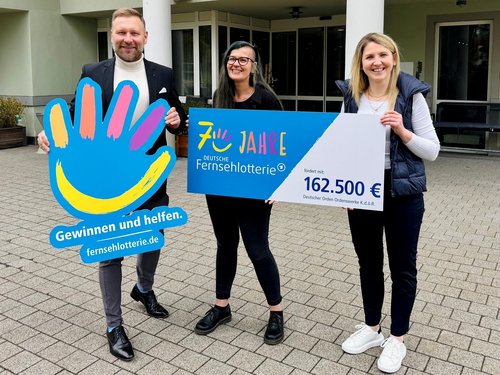 Drei lächelnde Personen halten große Schilder. Ein Mann zeigt das Logo der Deutschen Fernsehlotterie. Zwei Frauen präsentieren einen Spendenscheck über 162.500 Euro für den Deutschen Orden Ordenswerke zum 7-jährigen Jubiläum der Lotterie.