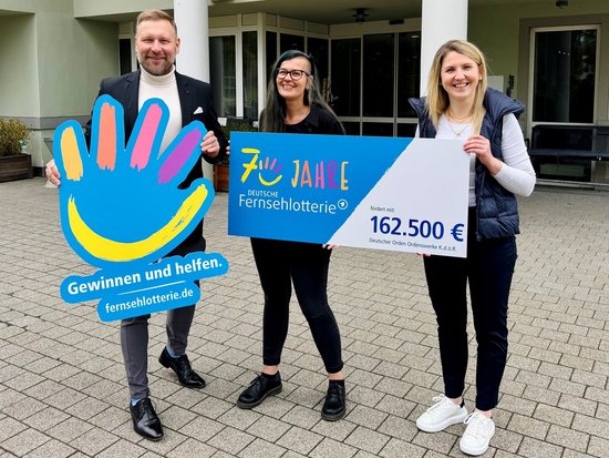 Drei lächelnde Personen halten große Schilder. Ein Mann zeigt das Logo der Deutschen Fernsehlotterie. Zwei Frauen präsentieren einen Spendenscheck über 162.500 Euro für den Deutschen Orden Ordenswerke zum 7-jährigen Jubiläum der Lotterie.