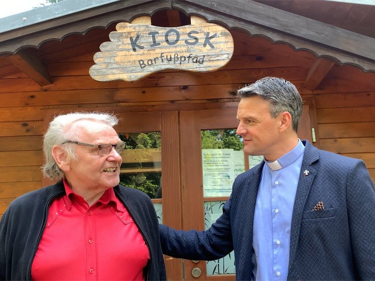 Pater Jörg vor dem Kiosk "Barfußpfad" hört einem älteren Mann zu