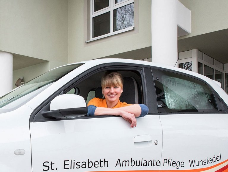eine junge Frau lächelt aus dem Fenster eines weissen Kleinwagens mit der Aufschrift: "St. Elisabeth Ambulante Pflege Wunsiedel"