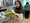 Eine lächelnde Frau mit schwarzem Hijab und hellem Oberteil hält eine große Schüssel mit grünem Salat in einer Küche. Im Hintergrund ist eine weitere Person bei der Arbeit an der Theke zu sehen.