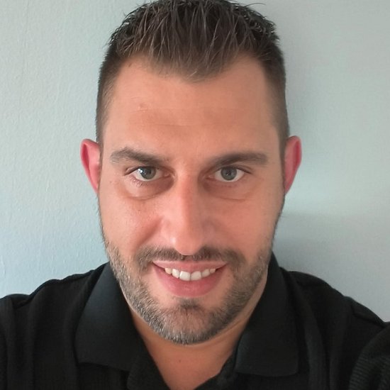 Leonardo Hohnjec Ein Mann mit kurzen dunklen Haaren und Bart lächelt direkt in die Kamera. Er trägt ein schwarzes Poloshirt vor einem hellblauen Hintergrund.